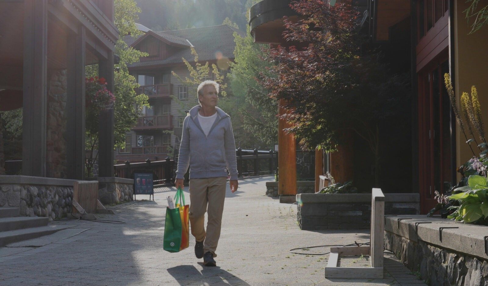 A person walking through a sunlit street with shops and buildings on a clear day, carrying a green reusable bag for their sustainable caregiving supplies.