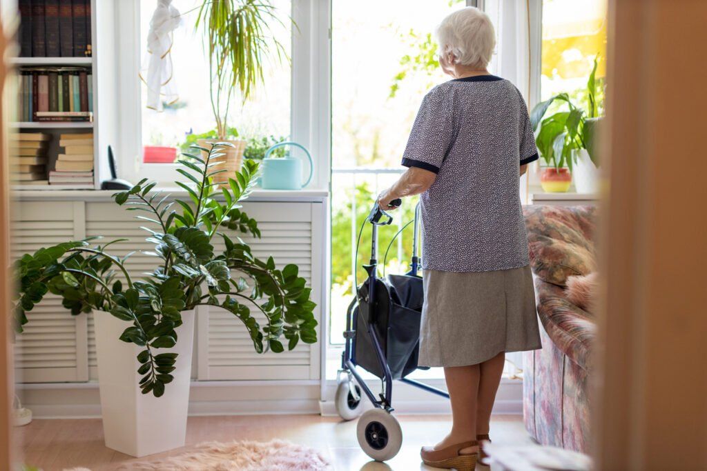 An older adult woman with dementia stands in front of her open house door with a walker.