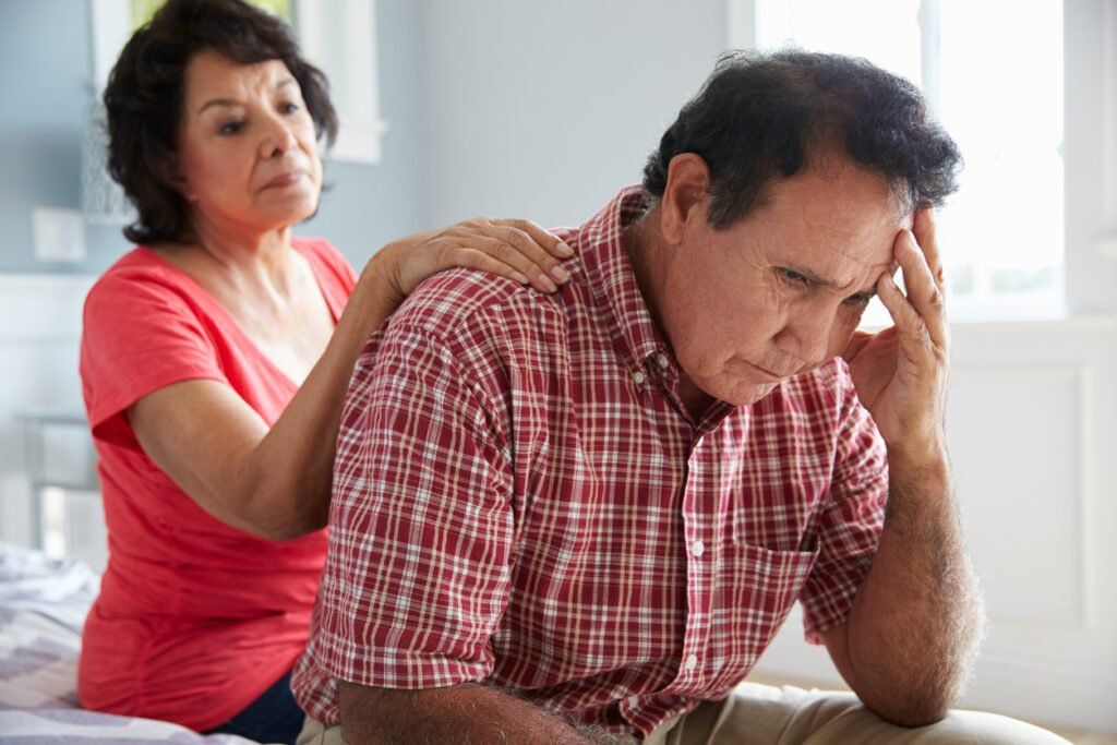 A man sits on the edge of a bed, resting his head on his hand with a distressed expression, while a woman places a comforting hand on his shoulder.