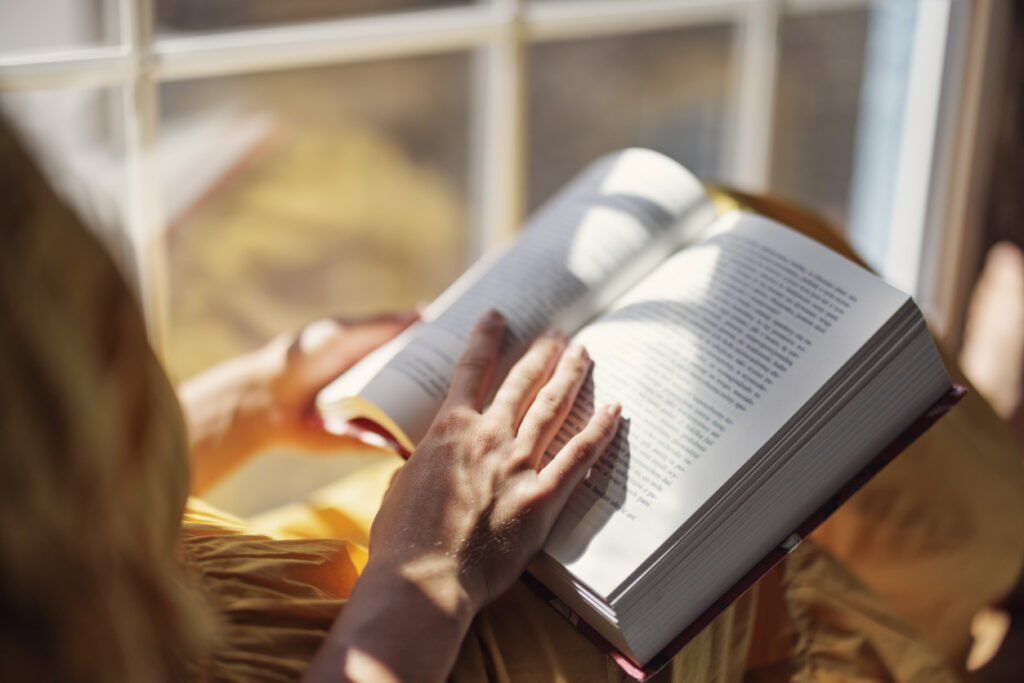 A person wearing a yellow garment is holding an open book while sitting near a window. Sunlight streams through the window, casting shadows on their hands and the book's pages. The background is blurred.