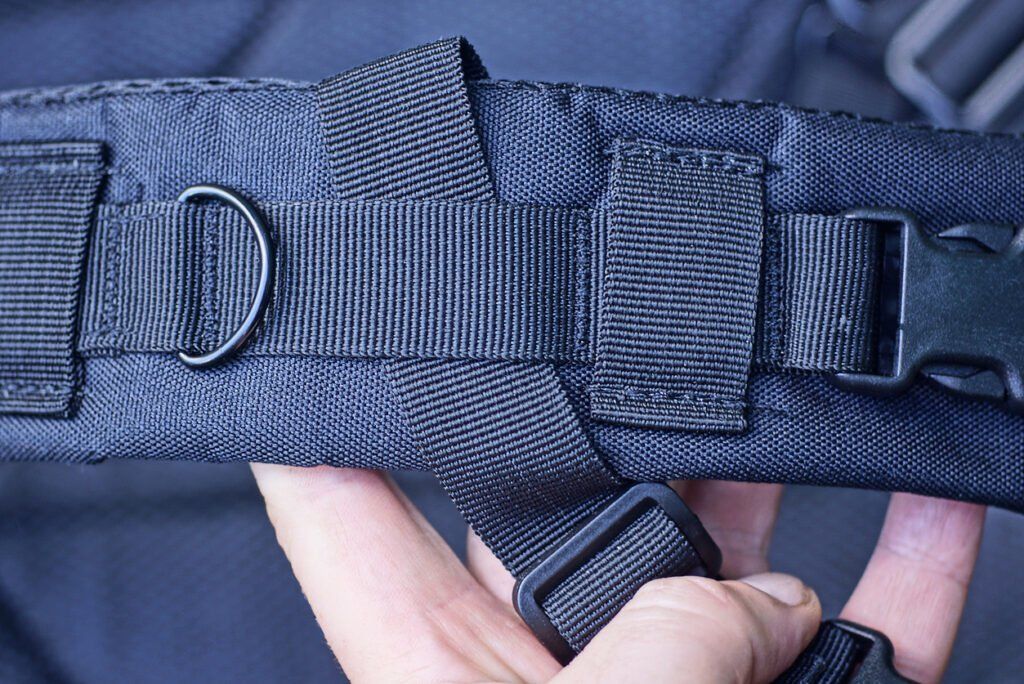 Close-up of a black gait belt with adjustable straps, a plastic buckle, and a metal D-ring, held by a person's hand.
