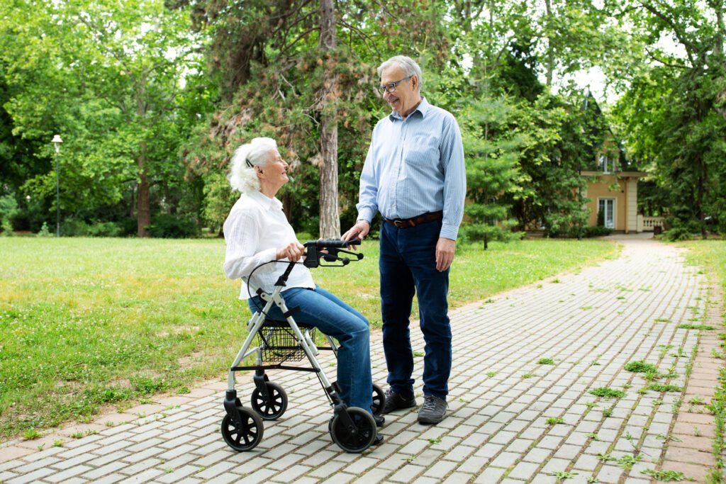 An older adult using a rollator walker sits on a paved path in a park while talking to another older adult standing nearby. The setting is outdoors with green trees, grass, and a house visible in the background.