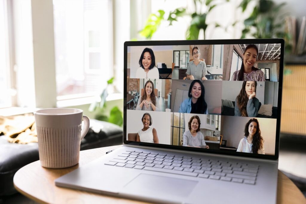 A laptop screen displays a virtual caregiver support group meeting with eight participants, all smiling and engaged in conversation. The laptop is placed on a wooden table next to a coffee mug. The background includes large windows with natural light, plants, and bookshelves, creating a cozy indoor setting.