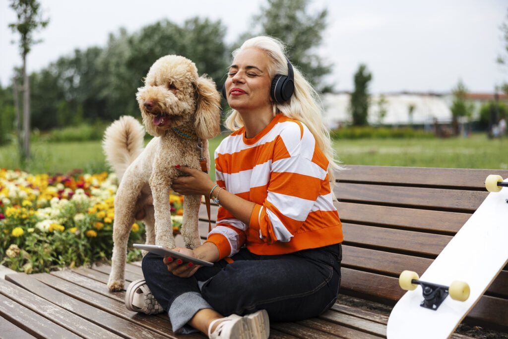 A woman with long blonde hair sits cross-legged on a wooden bench in a park, wearing black headphones and an orange-and-white striped sweater. She holds a tablet in one hand and pets a curly-haired dog standing beside her. A skateboard rests on the bench near her, and colorful flowers and greenery are visible in the background.