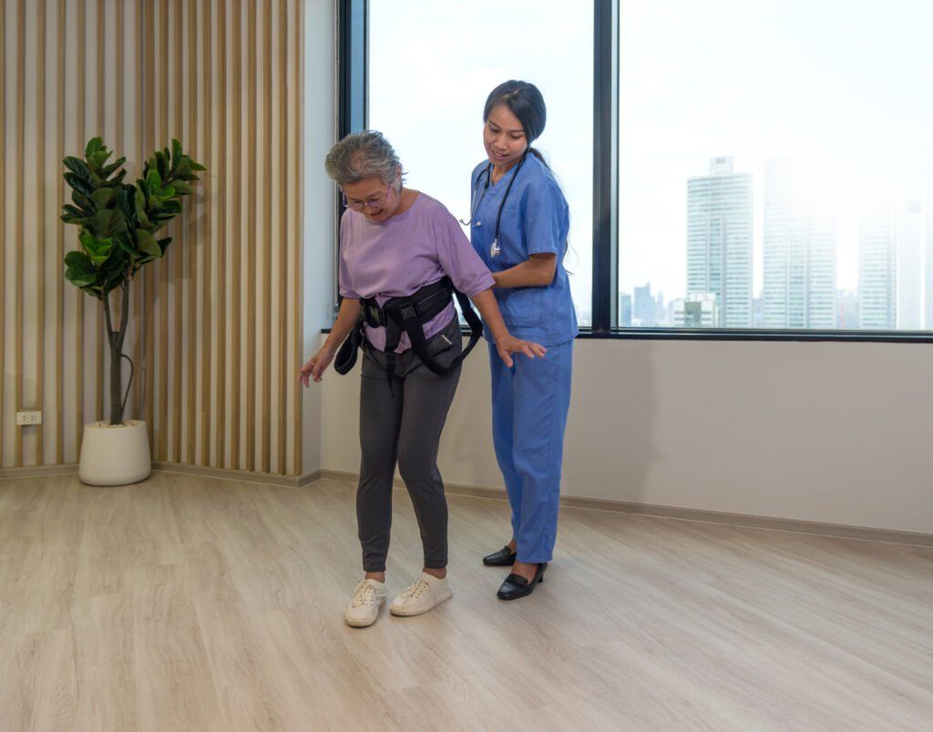 A healthcare professional in blue scrubs assisting an older adult wearing a purple shirt and a black gait belt during a physical therapy or mobility training session in a modern room with large windows and a wooden floor.