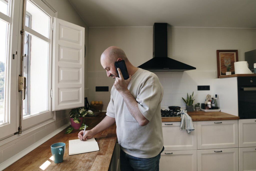 A man stands in a bright kitchen, holding a phone to his ear with one hand while jotting down notes in a notebook with the other. He is positioned near a countertop adorned with a mug, a potted plant, and other kitchen items, with natural light streaming in through an open window. The relaxed yet focused expression on his face suggests he is engaged in a productive conversation or task.