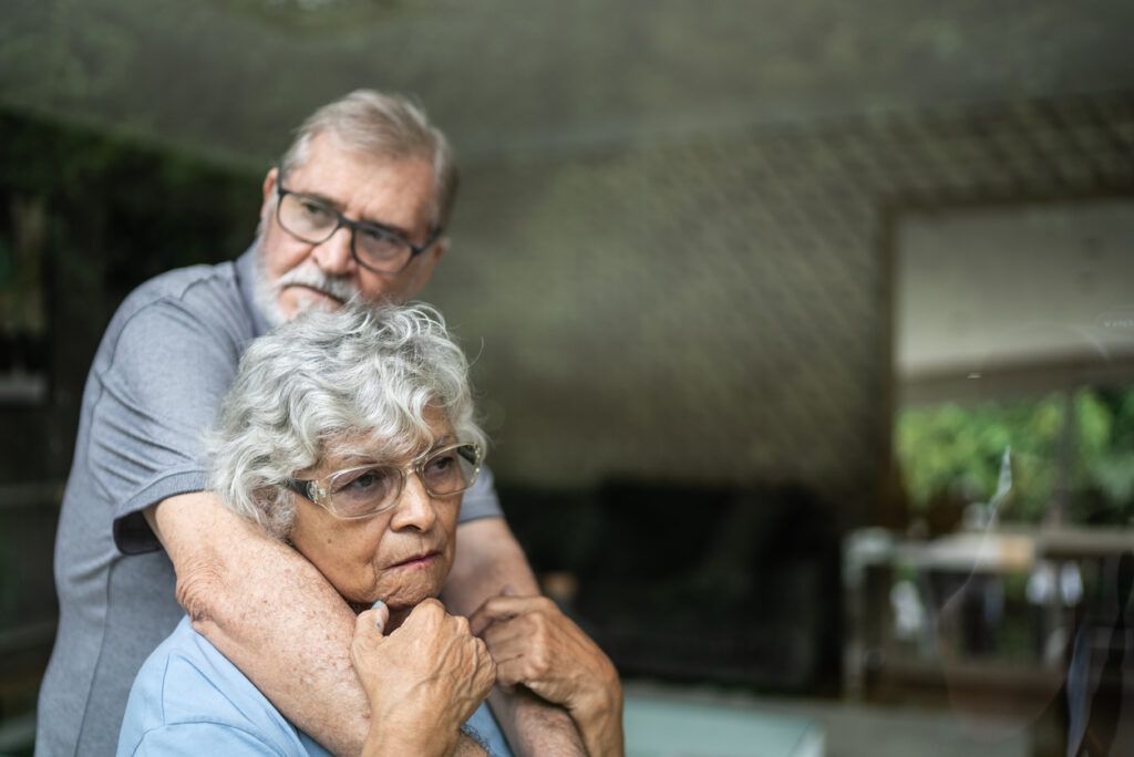 An older couple is standing near a window. The man, wearing glasses and a gray shirt, embraces the woman from behind with his arms wrapped around her shoulders. The woman, wearing glasses and a light blue shirt, looks thoughtful as she rests her hands on his arms. The window reflection shows greenery and a part of their living space.