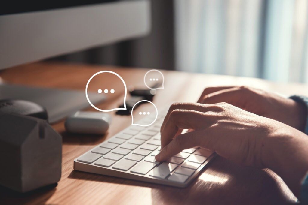 A close-up of hands typing on a white keyboard on a wooden desk. A desktop computer and other electronic accessories are visible in the background. Digital speech bubble icons with ellipses are overlaid on the image, representing online communication, messaging, or forums.