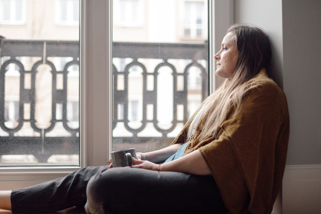 A woman with long brown hair sits on the floor by a large window, gazing outside with a thoughtful expression. She is wrapped in a brown shawl and holds a gray mug in her hands. The window overlooks a balcony with black wrought iron railings, and buildings are visible in the background.