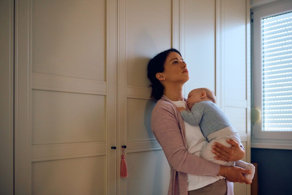 A woman stands against a light-colored wardrobe, holding a sleeping infant dressed in a light gray outfit. Natural light enters through a nearby window with closed blinds.