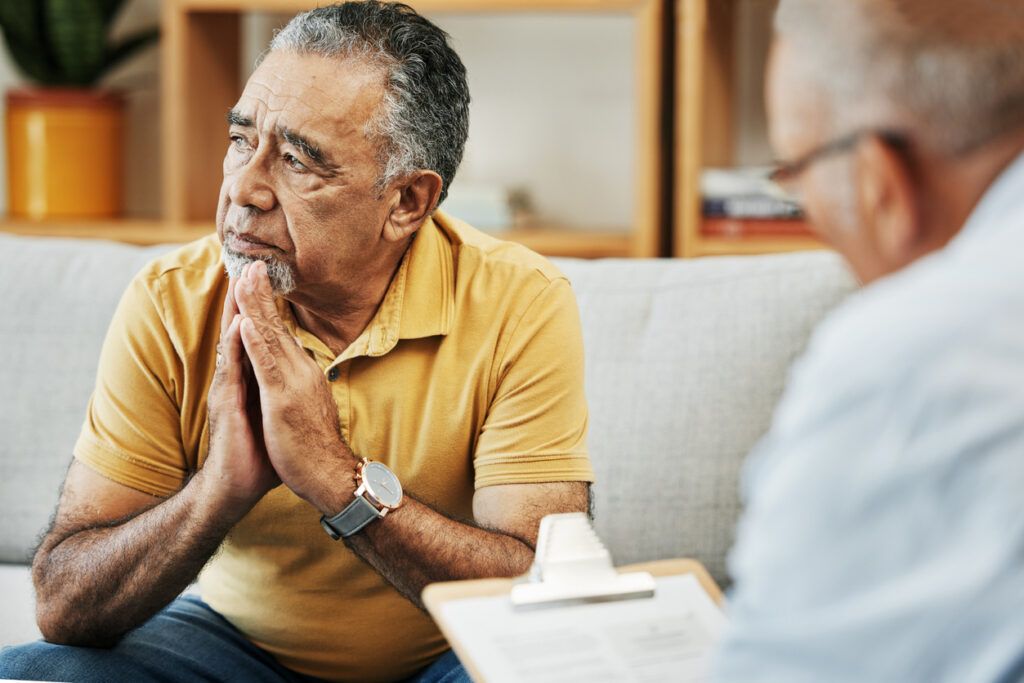 An older man with short graying hair, wearing a yellow polo sits on a couch with his hands clasped near his chin, looking thoughtful while speaking to a grief counselor who is partially visible in the foreground, holding a clipboard.