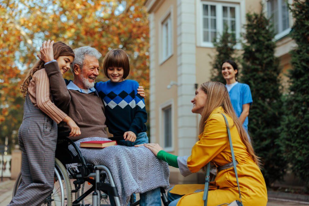 An elderly man with gray hair and a beard sits in a wheelchair, covered with a striped blanket, smiling warmly as two children, a boy in a blue sweater with an argyle pattern and a girl in overalls, hug him affectionately. A woman in a bright yellow coat kneels beside him, holding his hand and engaging in conversation. In the background, a caregiver in a blue uniform smiles as she observes the heartwarming interaction. The setting is outdoors, with autumn foliage and a beige building.
