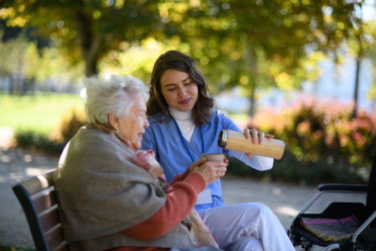 Elderly woman enjoying cup of coffee with her nurse in the park during warm autumn day. Young caregiver spending time with senior woman.