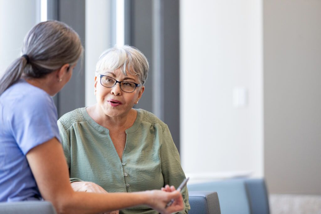 A caregiver is speaking to a healthcare worker who is wearing a light blue medical uniform. They are in a waiting area. The background consists of a neutral-colored interior with chairs and large windows.