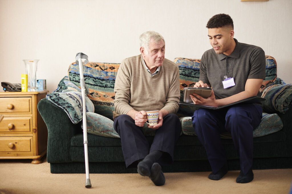 An older man with short gray hair, wearing a beige sweater and dark pants, sits on a patterned couch holding a mug. A crutch leans against the couch beside him. A younger man in a dark polo shirt and navy pants, wearing a name badge, sits next to him holding a tablet and showing him something on the screen. The background includes a wooden side table with a glass vase and a telephone.