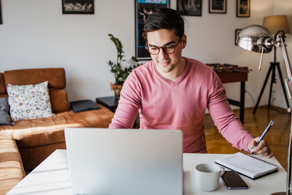 A man with short dark hair and glasses, wearing a pink sweater, is sitting at a white desk using a laptop. He is holding a pen and writing in a notebook. The background features a brown couch with decorative pillows, a floor lamp, framed pictures on the wall, and a foosball table.