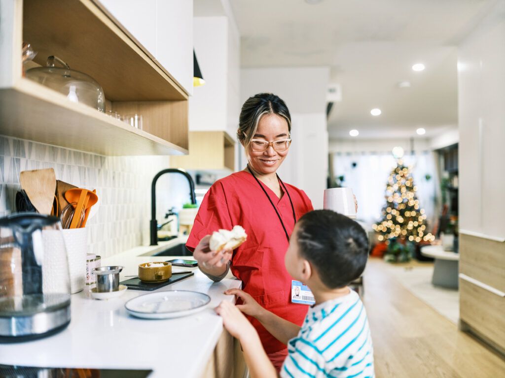 A woman wearing red medical scrubs and glasses stands in a kitchen holding a white mug and a piece of bread while smiling at a young boy. The boy, wearing a striped shirt, looks up at her. The kitchen has a white countertop, a black sink faucet, and wooden shelves with kitchen utensils and containers.