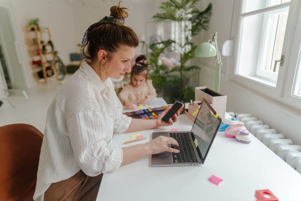 A woman with her hair tied up, wearing a white blouse, is sitting at a desk typing on a laptop while holding a smartphone. A young girl in the background is engaged in a creative activity with colored markers. The room is well-lit, with plants and various stationery items on the desk.