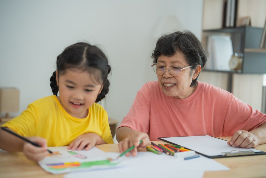 A young girl wearing a yellow t-shirt is drawing with colored pencils on a sheet of paper while sitting at a wooden table. Beside her, an elderly woman in glasses and a pink t-shirt smiles while assisting with the drawing. Colored pencils and a clipboard are scattered on the table, with a bookshelf and decorative items in the background.