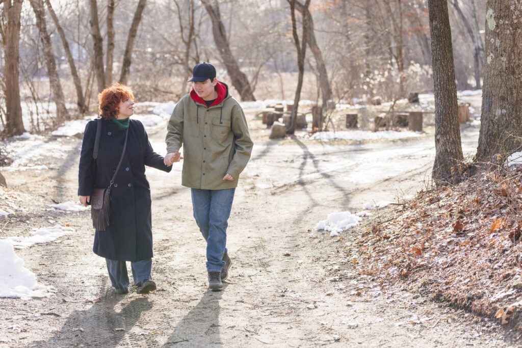 Two people walking hand-in-hand along a snowy forest path during winter, with bare trees in the background.