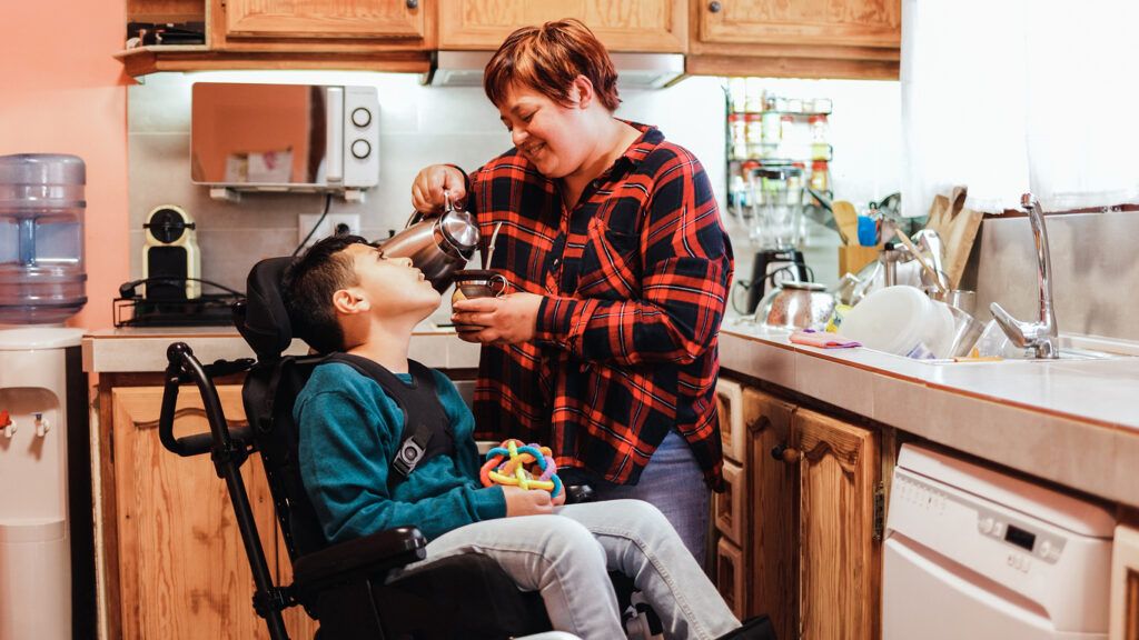 A boy with cerebral palsy in a wheelchair with a headrest and black frame is sitting in a kitchen, wearing a teal sweater and jeans. He is holding a colorful sensory toy in his lap. A hispanic woman in a red and black plaid shirt is pouring a drink from a metal pitcher into a cup while smiling at him. The kitchen has wooden cabinets, a microwave, a blender, and a dish rack.