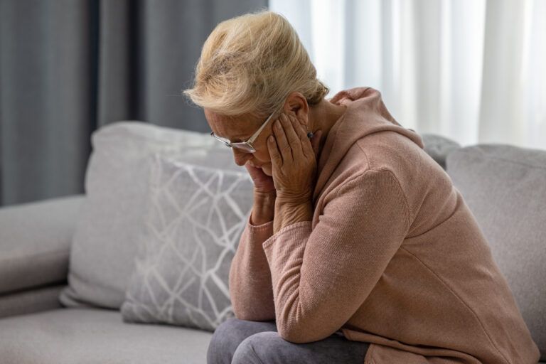 Distressed or depressed senior woman sitting alone on sofa at home holding her hand by the head. Loneliness concept
