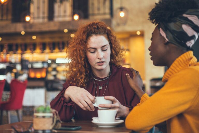 Two women sit at a café table, deep in conversation. One, a caring caregiver in a yellow sweater and headscarf, gently places a comforting hand on her friend’s arm as she sips her coffee.