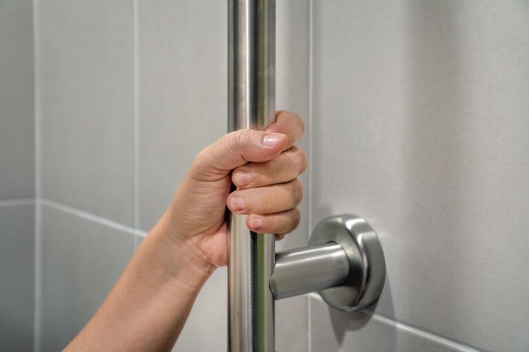 A close-up of a hand gripping a stainless steel grab bar mounted on a tiled wall, highlighting essential support for mobility in a bathroom or shower area.