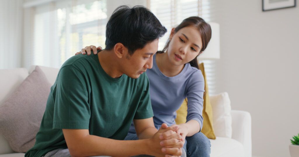 A woman sitting on a couch with her hand on the shoulder of a man beside her, who is leaning forward with his hands clasped.