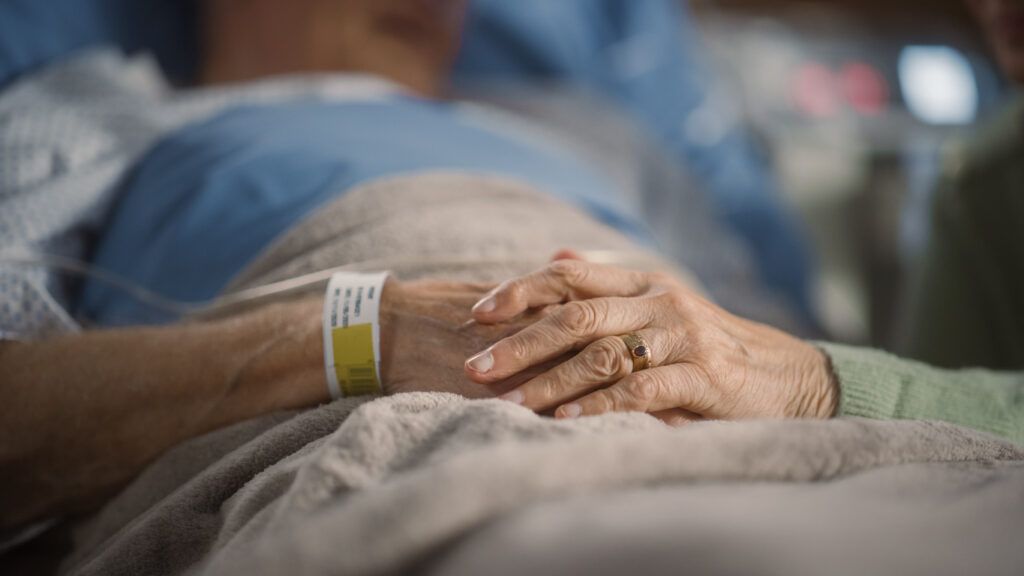 Close-up of two older adults holding hands, with one person wearing a hospital identification wristband and lying in a hospital bed covered with a blanket.