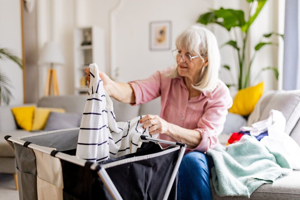 Older woman folding laundry in a living room with a laundry sorter nearby.