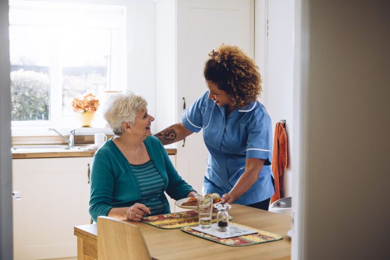 Caregiver serving a meal to a smiling elderly woman seated at a kitchen table.