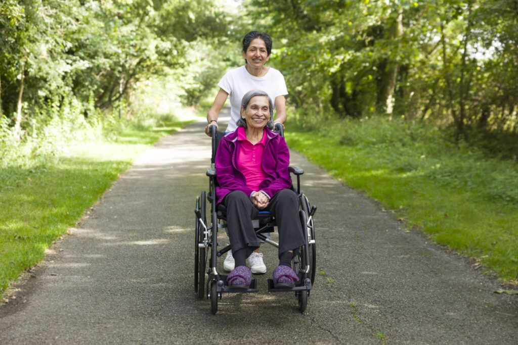Smiling caregiver pushing an elderly woman in a wheelchair along a forest path.