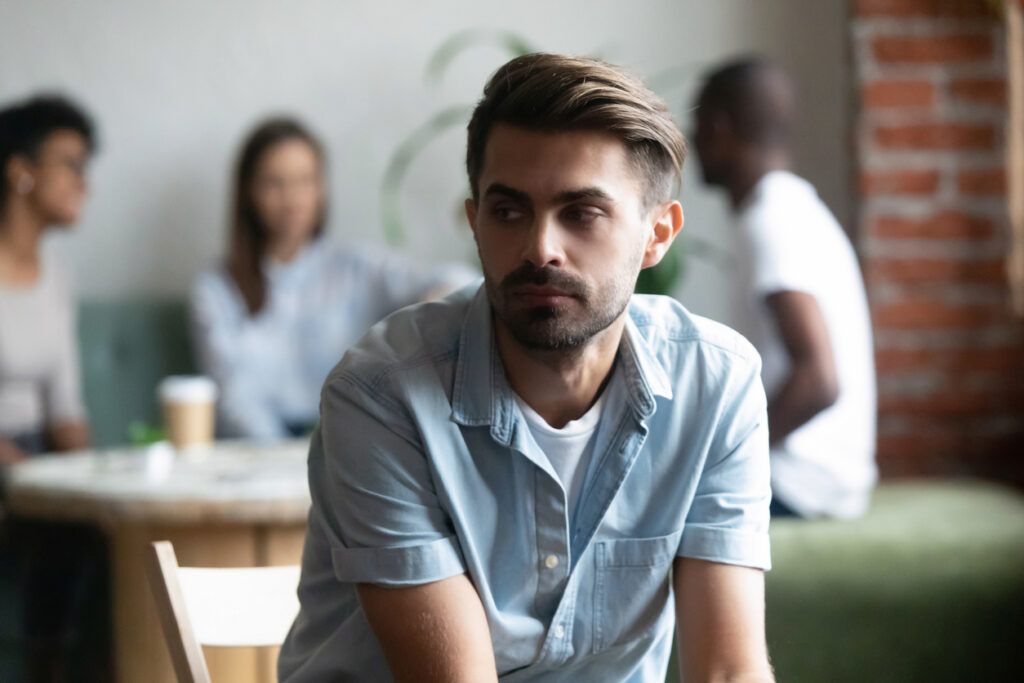 Man sitting alone in a group setting, looking away with a serious expression.