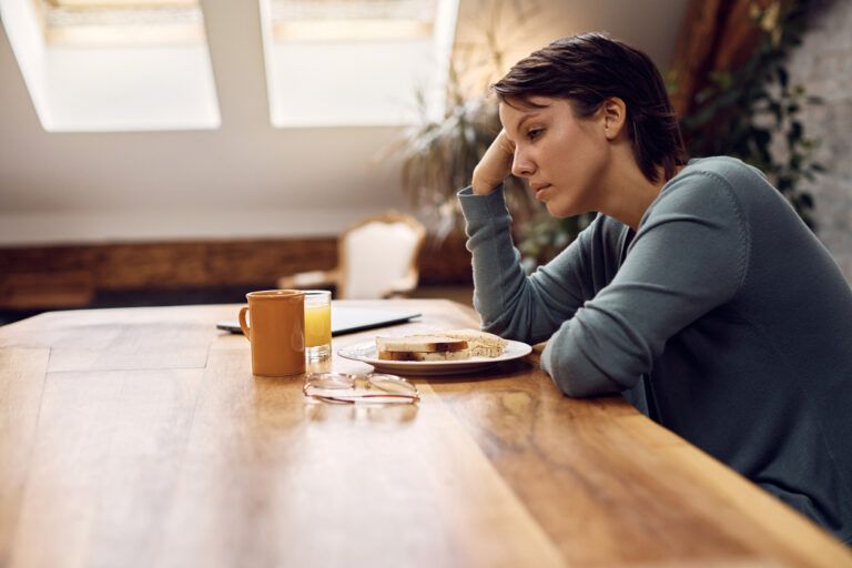 Woman sitting at a table with breakfast, resting her head on her hand in sadness.