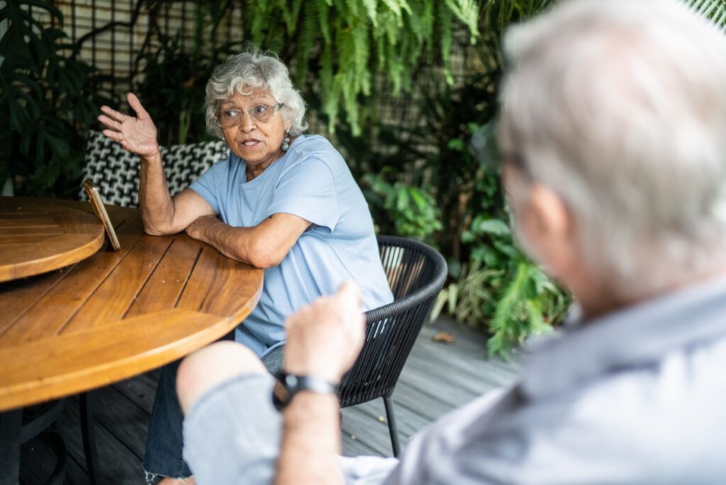 An elderly woman with gray curly hair is sitting at a wooden outdoor table, gesturing expressively with one hand while talking. She is facing an older man whose back is to the camera. Lush green plants and a casual patio setting surround them.