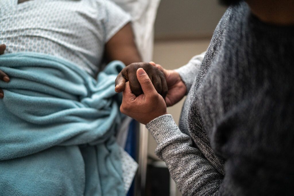A close-up of a person in a hospital bed covered with a blue blanket, while another person sits beside them holding their hand in a comforting gesture.