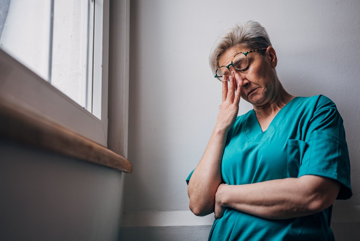 Healthcare worker pensive by the window, arm crossed.