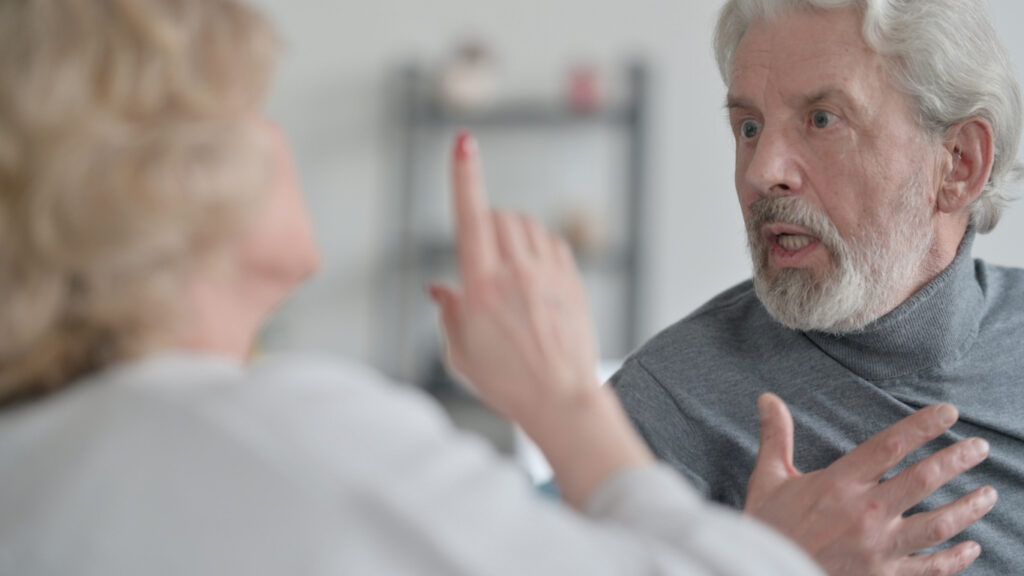 An older man gestures with his hand on his chest while speaking to a woman who is pointing her finger.