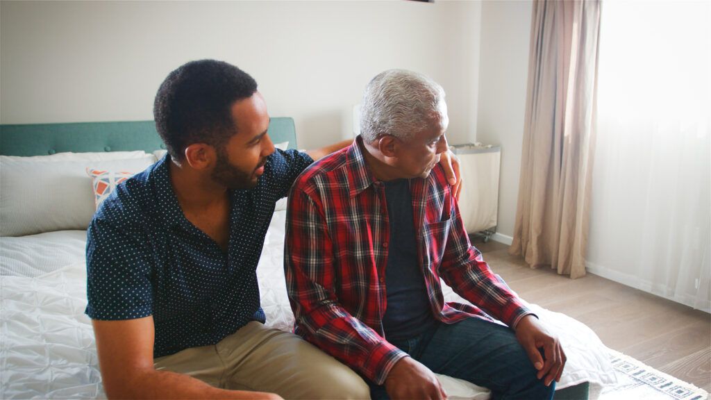 A younger man sits beside and gently supports an older man who is looking away while sitting on a bed.