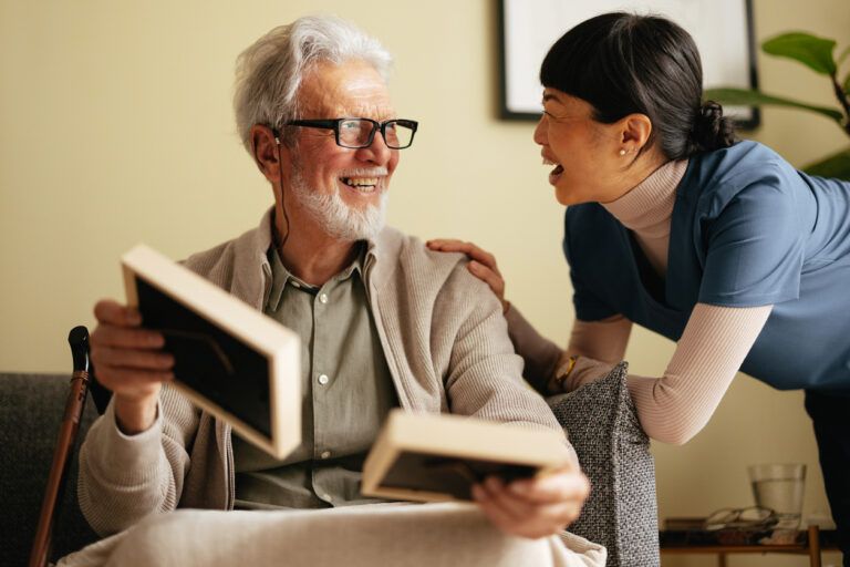 An older man with gray hair and glasses sits in a chair holding two photo frames, smiling at a woman standing next to him, who is also smiling. The woman, possibly his caregiver or nurse, supports him as he manages chronic kidney disease.