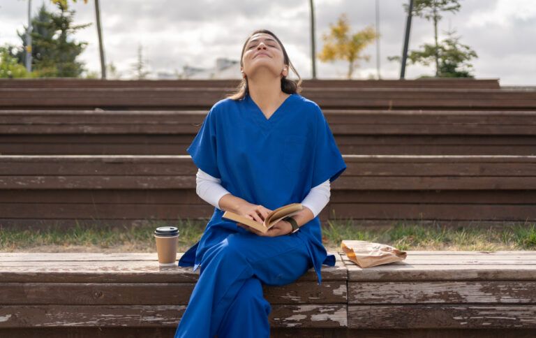 Caregiver sitting on bench outdoors with a book, coffee, and a paper bag, looking up.