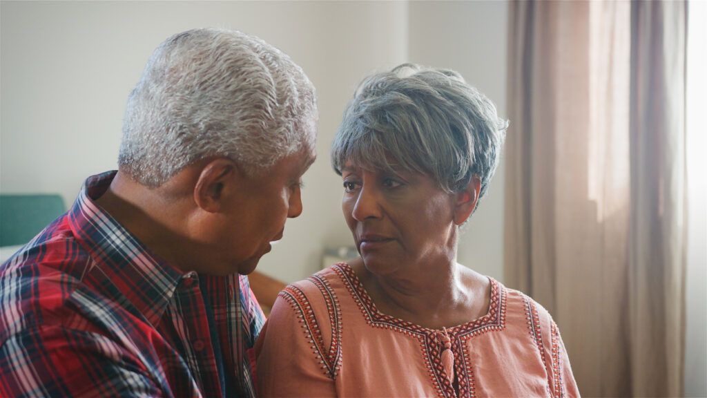 An older man and woman with dementia sit close together indoors, facing each other and making eye contact.