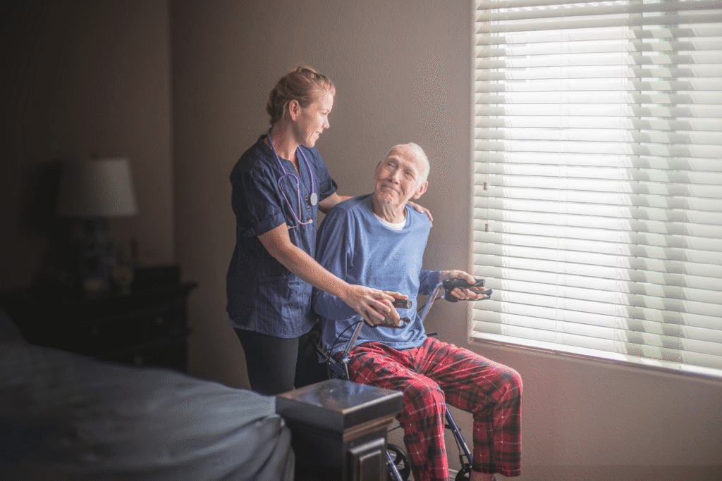 Nurse assisting elderly man in a wheelchair with a warm smile near window light