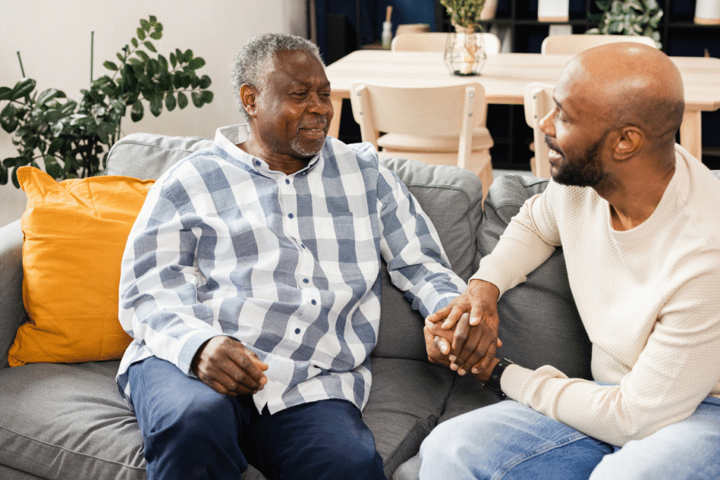 A middle-aged man smiles and holds hands with an older man while sitting together on a couch in a home setting.