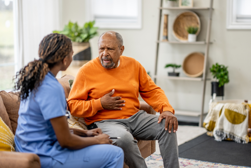 A stroke survivor wearing an orange sweater sits on a couch and talks to a home nurse wearing blue scrubs.