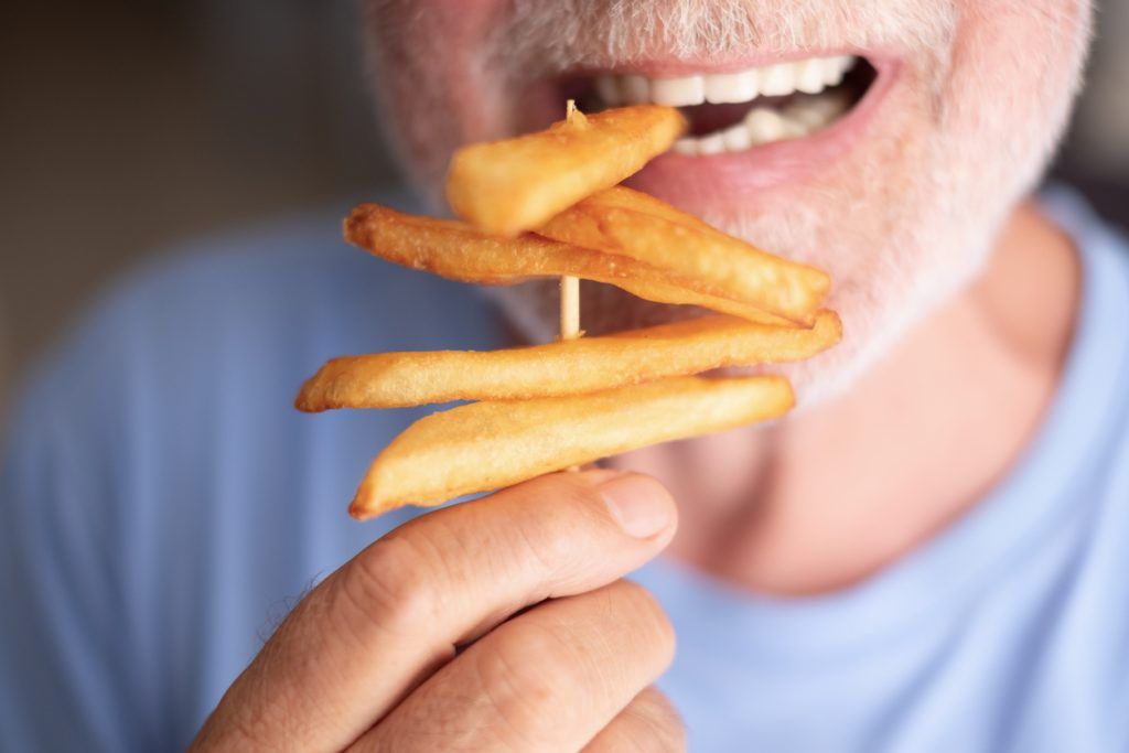 Close-up of an older man holding a skewer with several French fries, about to eat one.
