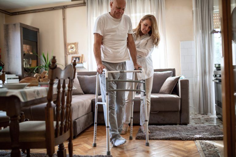 Caregiver assisting an older man using a walker inside a home living room setting.