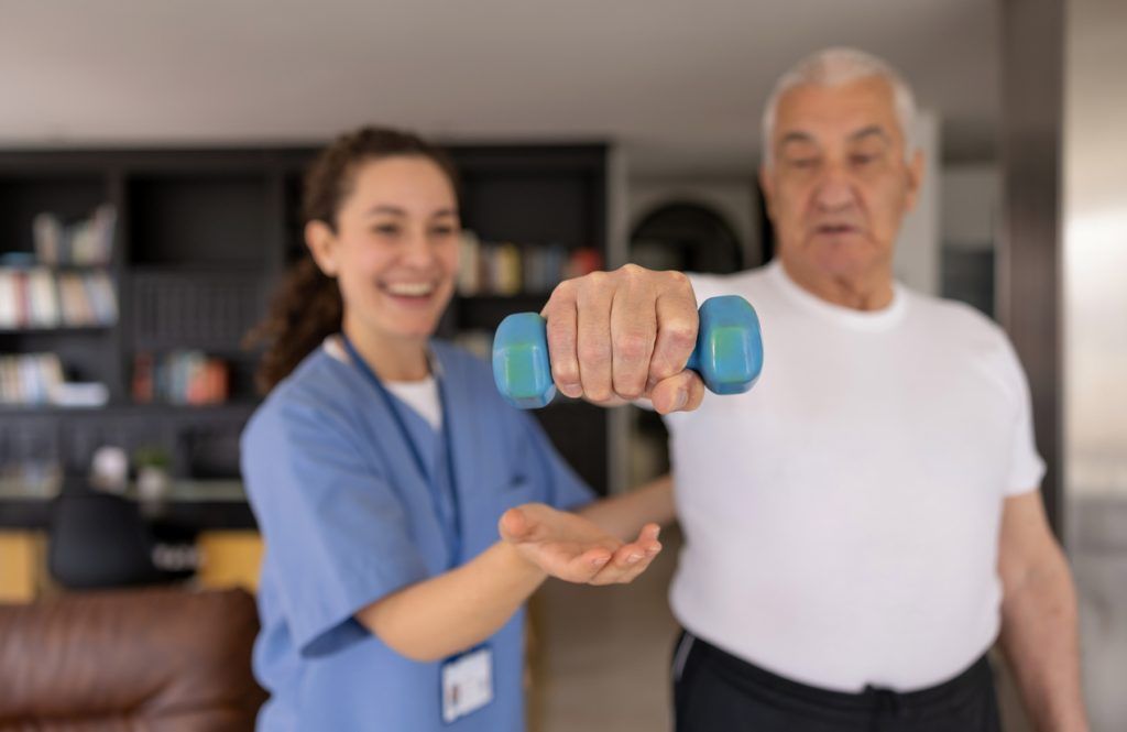 Caregiver assisting a senior man lifting a dumbbell in an exercise routine.