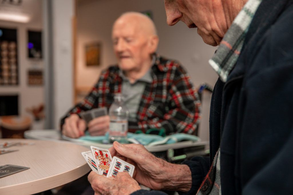 Two older adult men sitting at a table holding playing cards in their hands.
